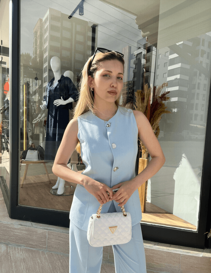 Bebe Mavisi Düğmeli Yelekli Pantolon Takım giyen beyaz çantalı, sarı saçlı kadın.